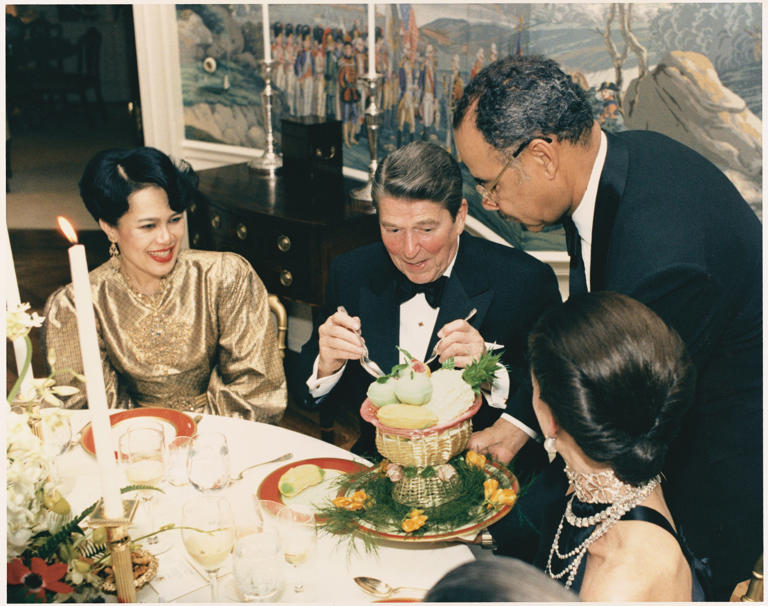 This is a photograph of President Ronald Reagan taking a scoop of sorbet from a pulled-sugar basket as Queen Sirikit, queen consort of Thailand, looks on. The Reagans honored her with an intimate black tie dinner on March 11, 1985.