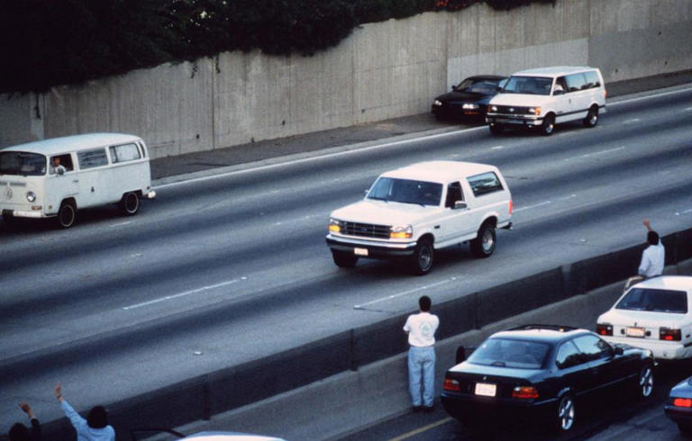 Motorists wave as police cars pursue the Ford Bronco (white, R) driven by Al Cowlings, carrying fugitive murder suspect O.J. Simpson, on a 90-minute slow-speed car chase June 17, 1994 on the 405 freeway in Los Angeles.