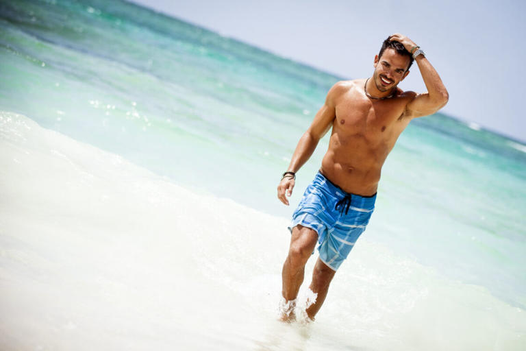 An attractive young gay man wearing swim trunks on the beach by the ocean of an LGBTQ+-friendly Caribbean island