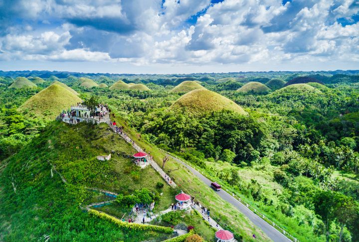 <p>The location has around 1,200 cone-shaped hills spread over 50 square kilometers. These hills are named “Chocolate Hills” because their green grass becomes brown during the dry season, making them look like chocolate kisses. This unique landscape is a popular tourist attraction as a designated UNESCO World Heritage Site.</p>