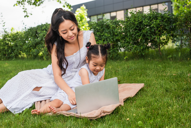 Mom and daughter sitting in the grass on a blanket working on laptop