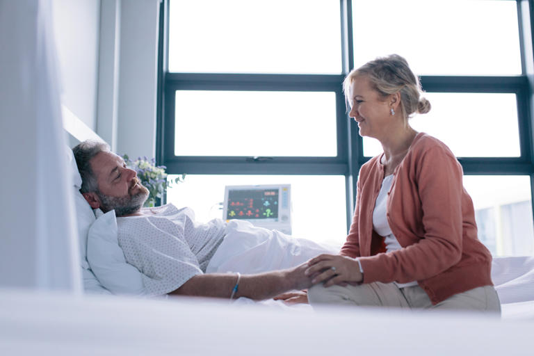  Female talking with male patient lying in hospital bed.