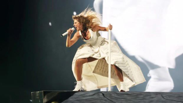 A woman in a white dress with book-style text printed on it leans forward with a fierce expression, holding a microphone on an elevated platform above a crowd. It's Taylor Swift, with light-toned skin and blonde hair.