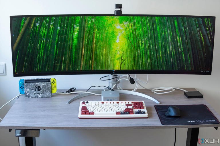 A desk setup with multiple devices, including a Geekom A8 PC under a large ultrawide monitor