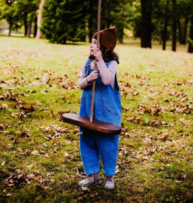 Child in a denim jumpsuit and hat plays on a tree swing