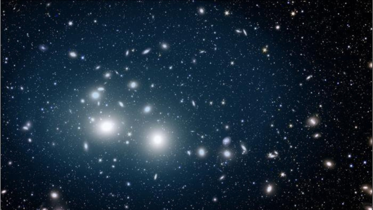 A Euclid image of the Perseus cluster of galaxies bathed in a gentle, soft blue light emanating from orphan stars. These orphan stars are dispersed throughout the cluster, extending up to 2 million light-years from its centre.