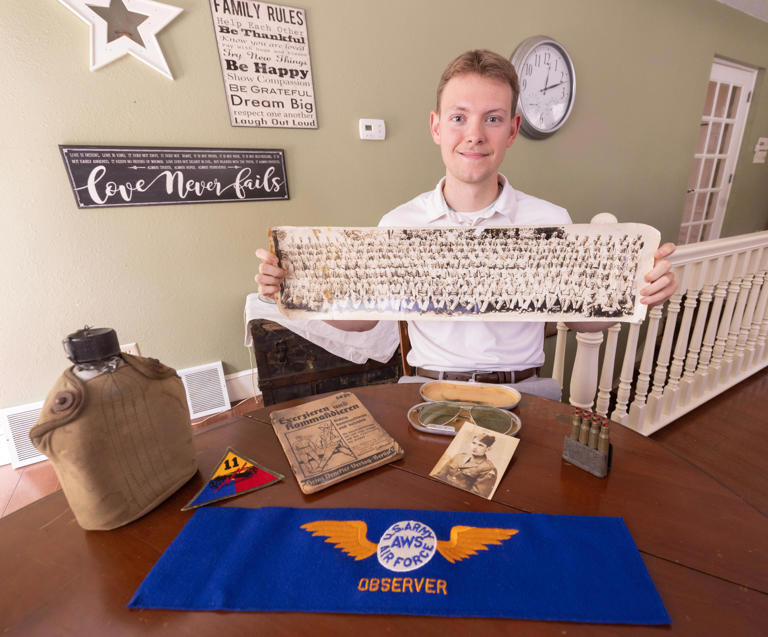 Thomas Fulk, a Kent State University at Stark history major, is traveling to Normandy, France, for the 80th anniversary of D-Day. He holds a photograph of his great-grandfather Russell Fulk in his Army basic training group photo while standing with some of his World War II artifacts at his East Sparta home.