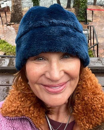 A woman wearing a blue hat and a brown jacket smiles for a selfie with a river and trees in the background