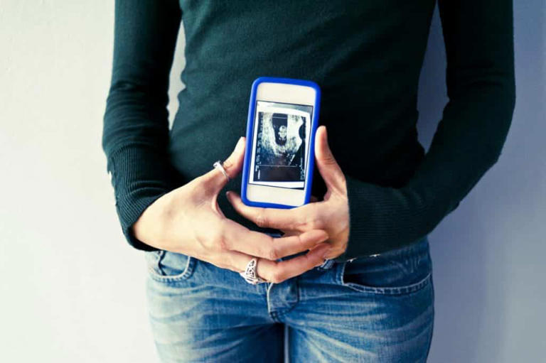 A person wearing a dark long-sleeve shirt and jeans is holding a smartphone with both hands, displaying an ultrasound image from the First Trimester.