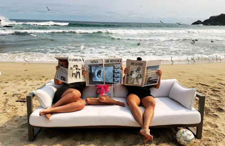 En una playa con el mar de fondo, tres personas están sentadas en un sofá blanco sobre la arena. Las dos adultas a los lados leen periódicos que dicen 'FAMILIA' y 'DUAYHE', mientras que la persona en el centro, aparentemente una niña, sostiene un periódico que dice 'NIÑO DE RIVERA' con una imagen de ultrasonido. A un lado del sofá, hay un balón de fútbol en la arena