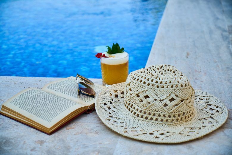 White sun hat, open book, and cocktail next to a blue swimming pool
