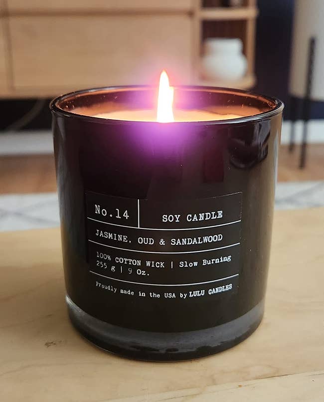 A lit candle with black glass and white type text saying it's a jasmine, oud, and sandalwood scent with a cotton wick