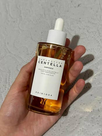 Hand holding a bottle of SKIN1004 Madagascar Centella Ampoule, 100 ml, on a textured white surface