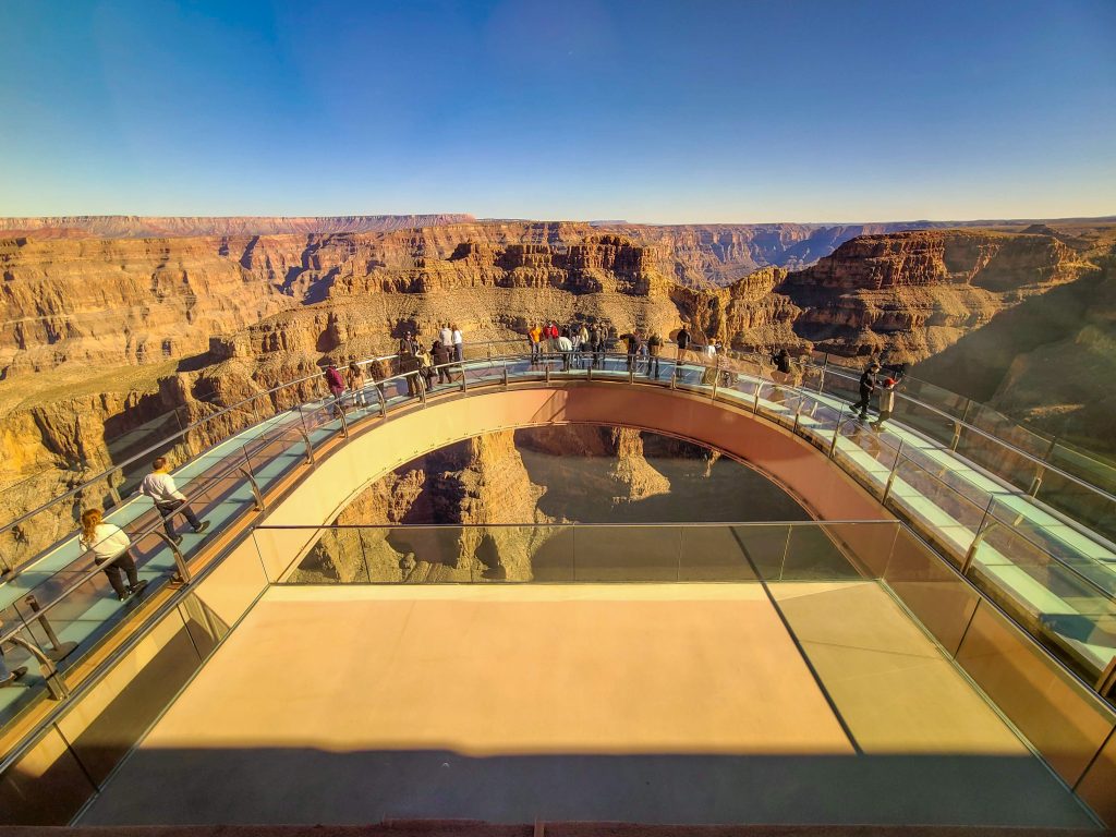 <p>The glass Skywalk at Grand Canyon West is a unique feature that distinguishes GCW from other parts of the canyon. It’s a 70′ loop extending over the rim with a translucent glass floor. Judging from the T-shirts at the gift shop, it triggers some people’s vertigo. However, we felt entirely secure on the platform without even one butterfly.</p> <p>The views, however, are outstanding. There’s a dad joke here about out-standing 1000′ above the canyon floor, but all kidding aside, they are amazing. I’ve never looked back onto the rim before, and it felt like I was flying over the canyon.</p> <p>Entrance to the Skywalk isn’t included with a general admission ticket, but you can purchase it separately or bundle it in with package deals. You almost have to purchase a Skywalk ticket just to experience the feeling and views.</p> <p>Additionally, you can’t take your own pictures, which is just as well considering the chaos that would ensue with selfie sticks and the stupid things people will do to ‘get one for the Gram.’ However, trained photographers are there to take shots that will amaze your friends and scare your kids (or parents). The photo package also includes some spectacular scene shots that we could only dream of taking.</p>