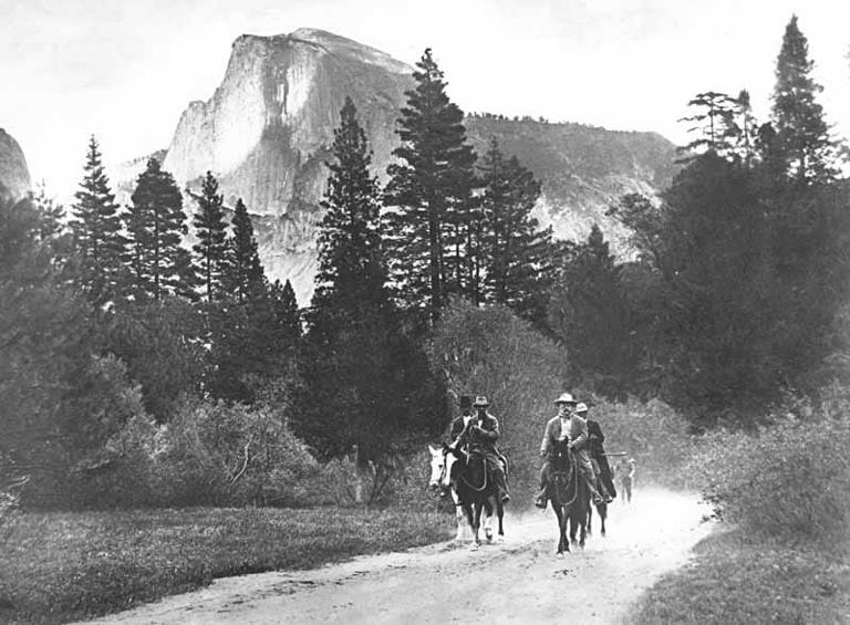 President Theodore Roosevelt and John Muir riding horses along a road in Yosemite Valley, with Half Dome in the distance, accompanied by Park Rangers Archie Leonard and Charles Leidig, followed by unidentified man on foot.