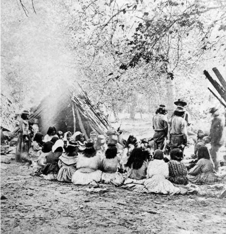 Group of about twenty-six Native Americans seated and standing beside a cedar bark structure, near the Merced River, Yosemite Valley, 1872.