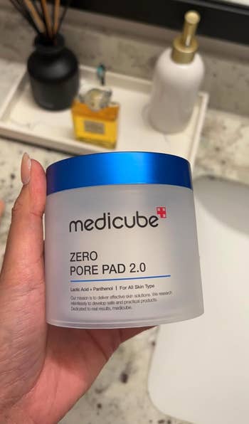 Hand holding Medicube Zero Pore Pad 2.0 container, a skincare product for treating pores. Background includes bathroom items
