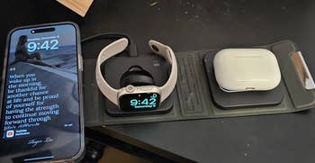 iPhone displaying a quote next to a smartwatch charging on a dock alongside a pair of wireless earbuds