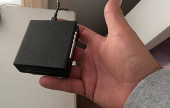 A person's hand holding a rectangular electronic device with a cord connected to it. The item is likely a tech gadget used for charging or data storage