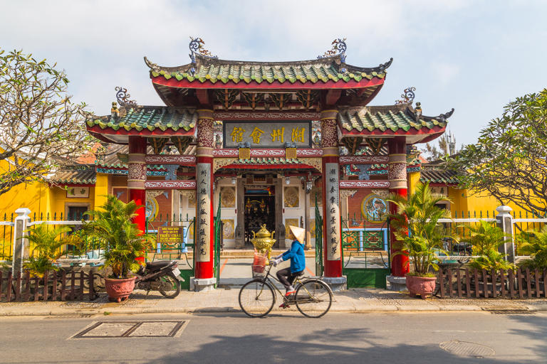 The outside of the Trieu Chau Assembly Hall in Hoi An, Vietnam during the day. A person with a traditional conical hat can be seen going past on a bike.