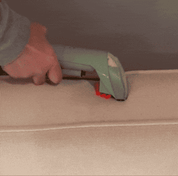 A hand uses a green handheld vacuum cleaner to clean the fabric of a beige sofa cushion