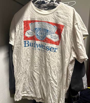 A white graphic T-shirt with a large Budweiser logo hangs on a clothes rack. The shirt is wrinkled and appears to be in a closet