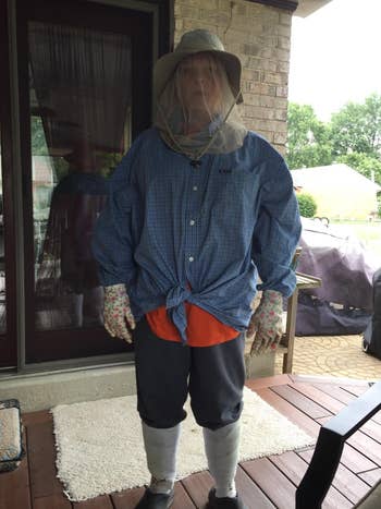 A person stands on a patio wearing protective clothing including a wide-brim hat with netting, a long-sleeve shirt, gloves, and long socks pulled over pants