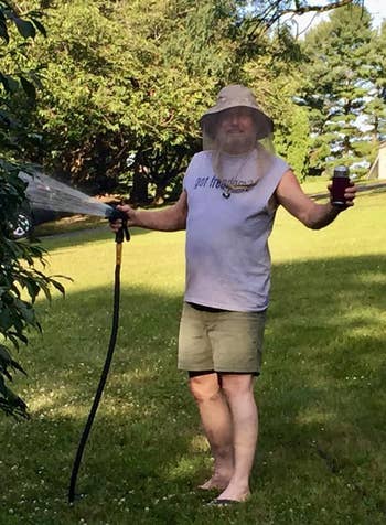 Man wearing a sun hat, sleeveless shirt, shorts, and a mesh face covering waters plants with a hose while holding a drink in a garden