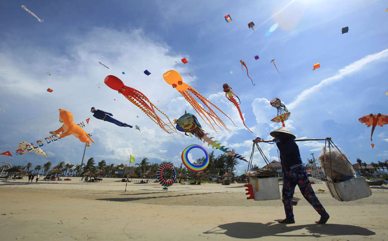 Slide 5 of 33: In this Sunday, June 11, 2017, photo, a food vender walks under flying kites on Tam Thanh beach during an International Kite Festival in Quang Nam province, Vietnam. Hundreds of flying giant sea creatures, animal shaped and folklore inspired kites from 2