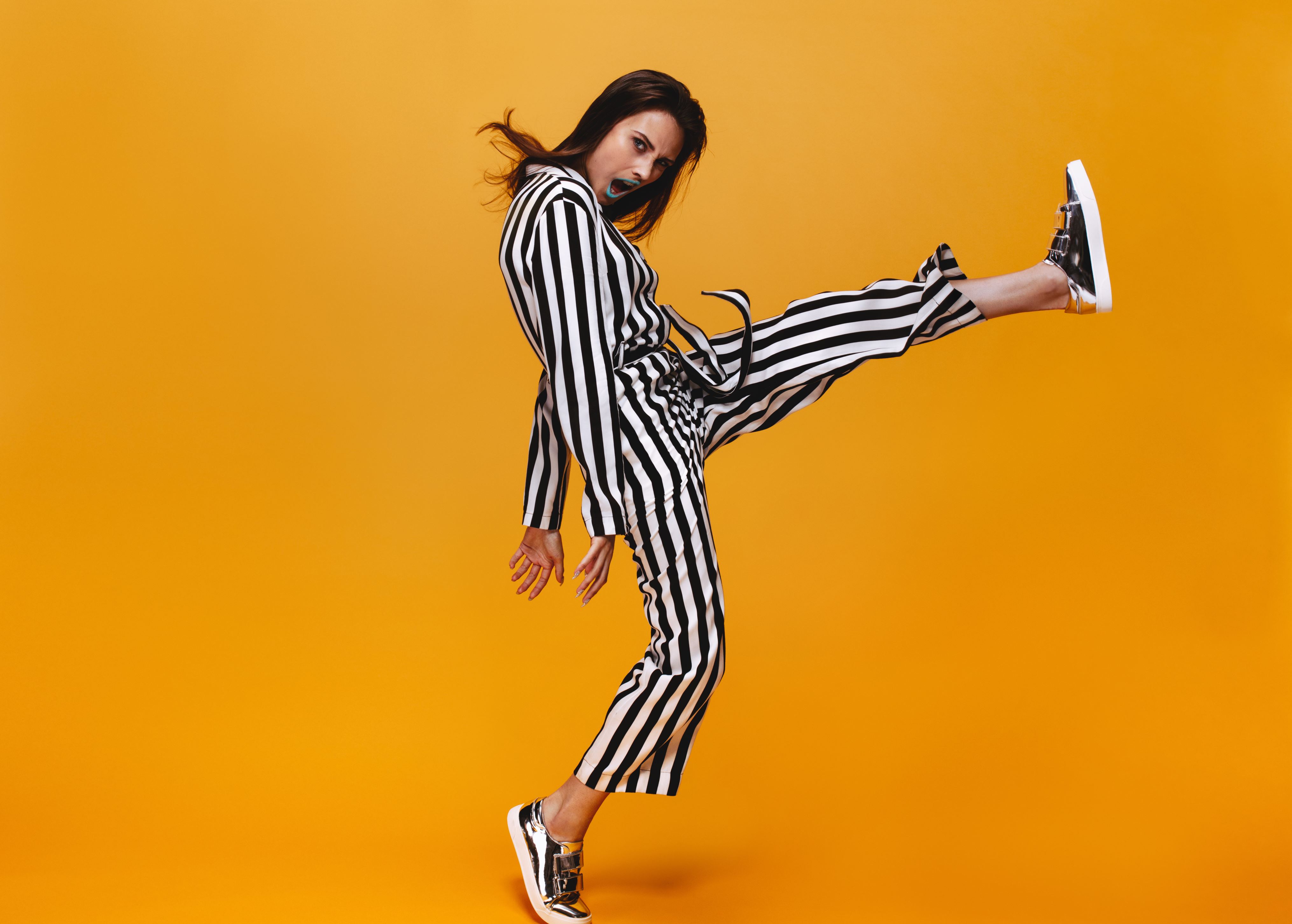Full length of young woman in jumpsuit kicking over orange background. Funky female with raised leg looking at camera.