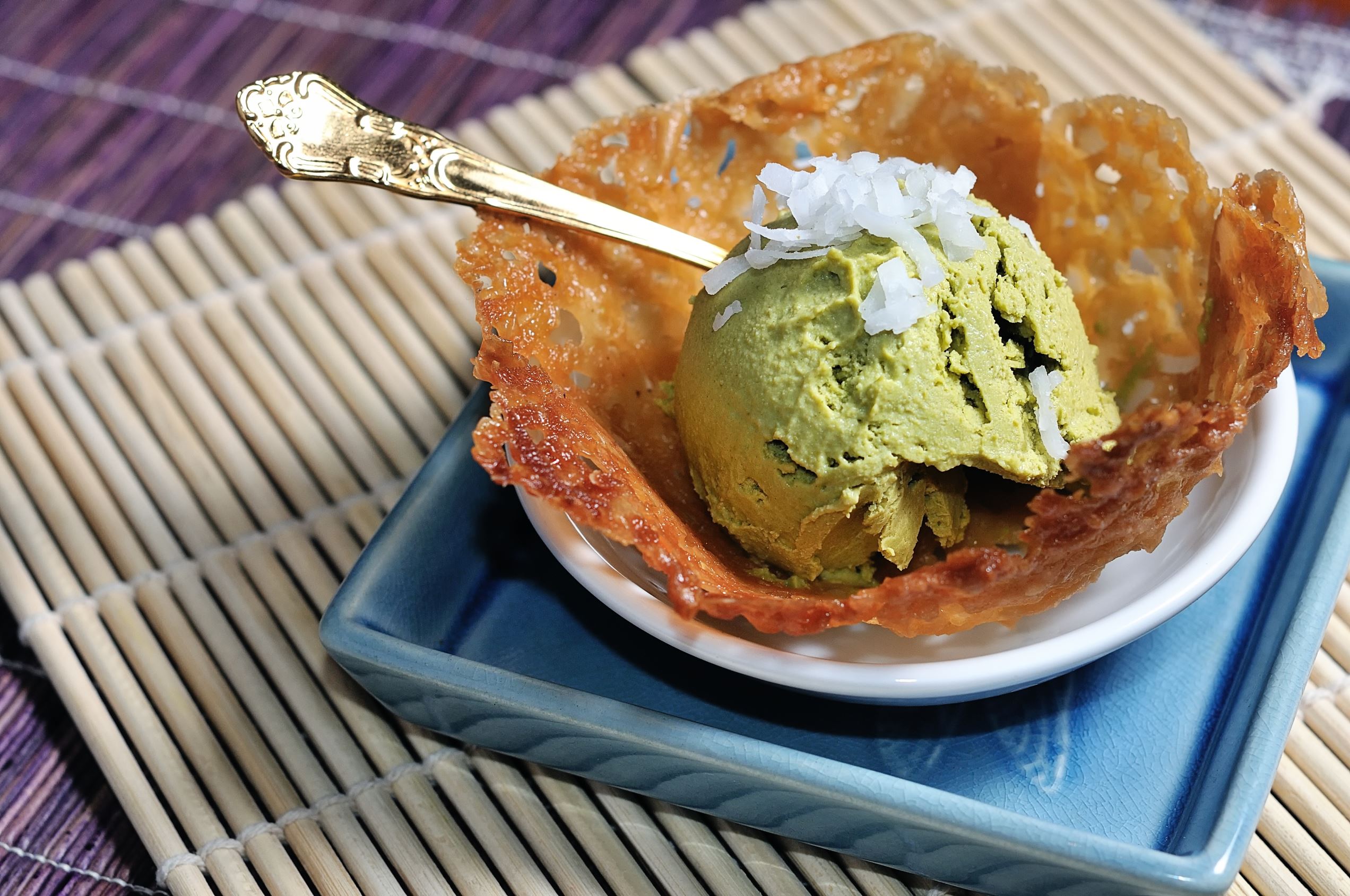 Slide 7 of 20: Matcha green tea ice cream in a coconut tuile cookie bowl, garnished with flakes of coconut.  Bamboo placemats in the background.